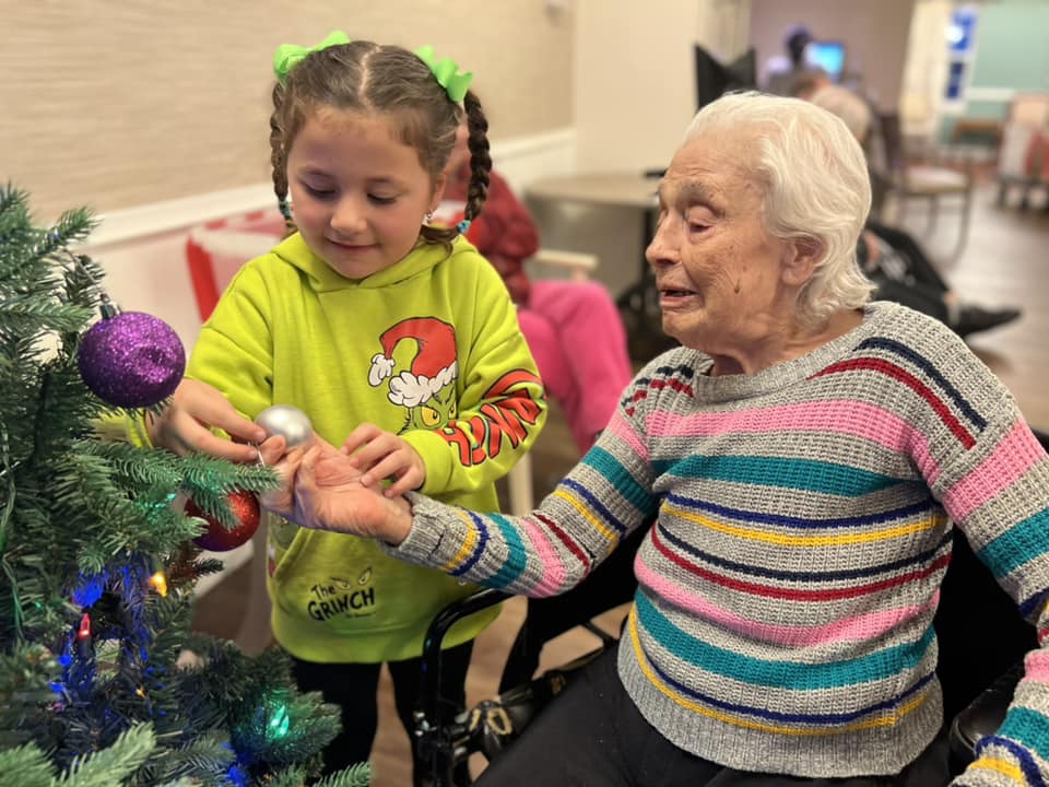 An elementary school-aged child helping an elderly woman hang ornaments on an evergreen tree