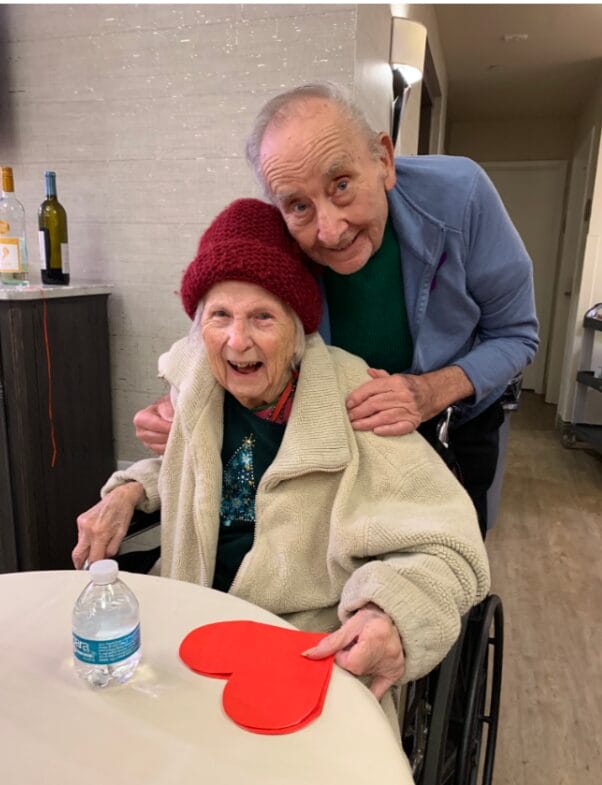 An elderly couple smiling with a cut-out of a heart on a table in an assisted living facility