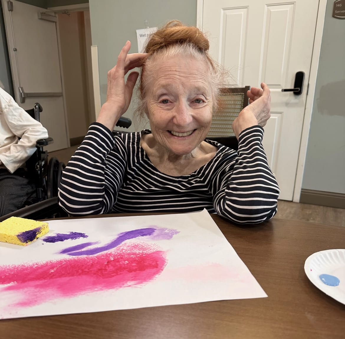 An assisted living resident smiling with artwork in front of her