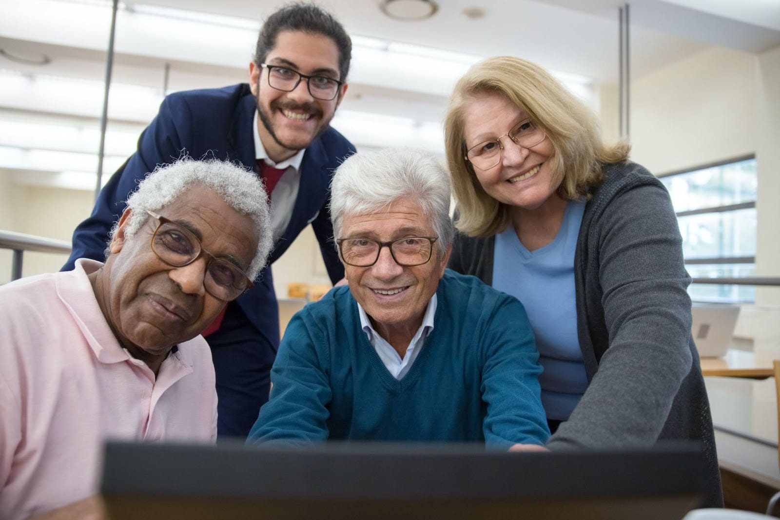 A group of seniors and a young man in a suit smiling behind a computer