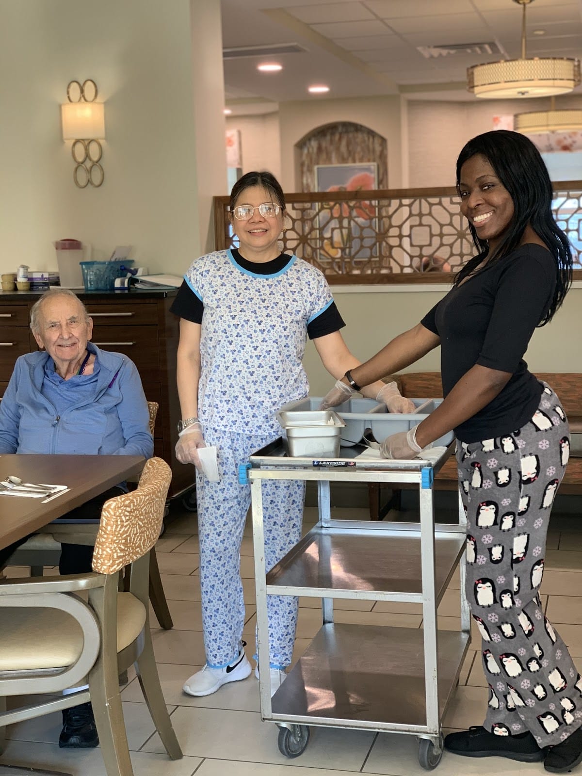 An elderly gentleman eating in an assisted living facility as two care workers stand by smiling