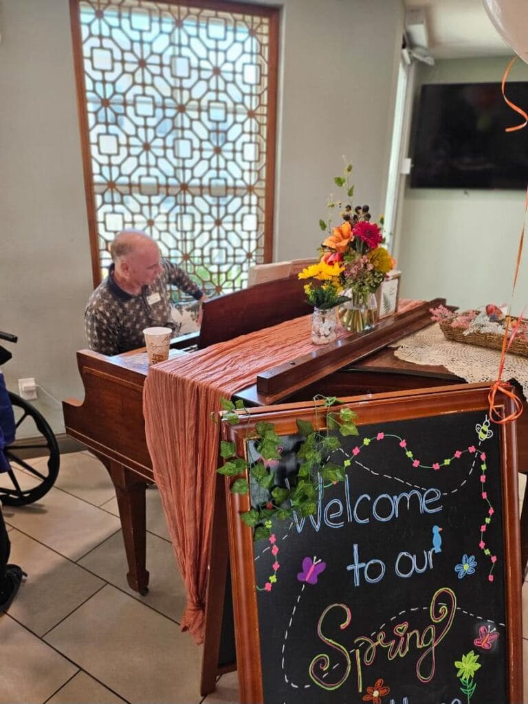 A picture of an elderly person playing piano in front of a sign that reads Spring Open House