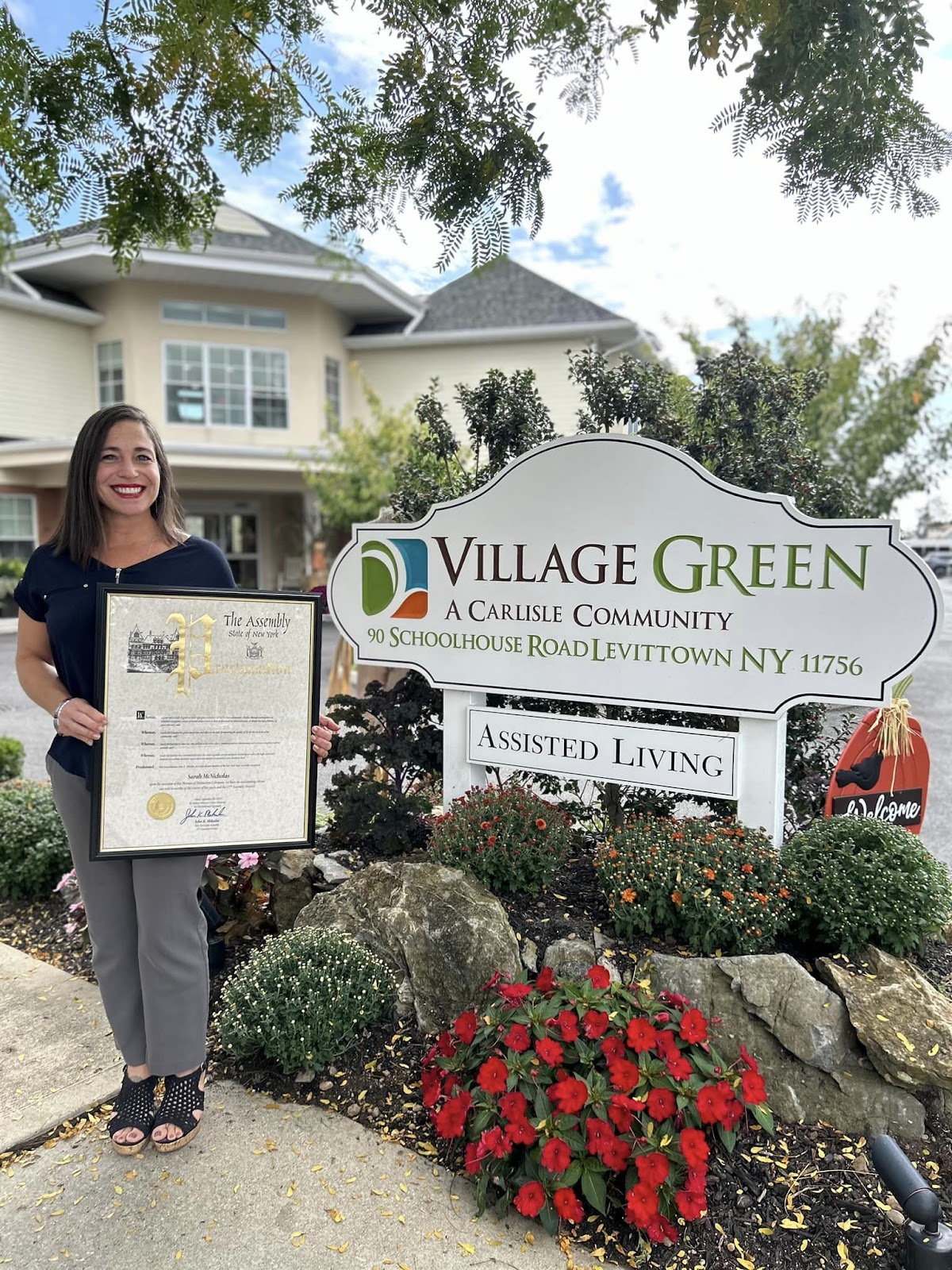 A Village Green caregiver smiling in front of the outdoor Village Green sign, all while holding a framed award