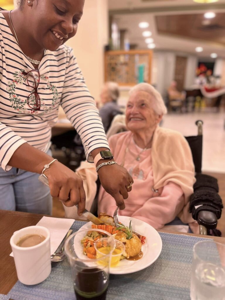 A picture of a smiling resident smiling at the staff member as the staff member cuts up food on a plate