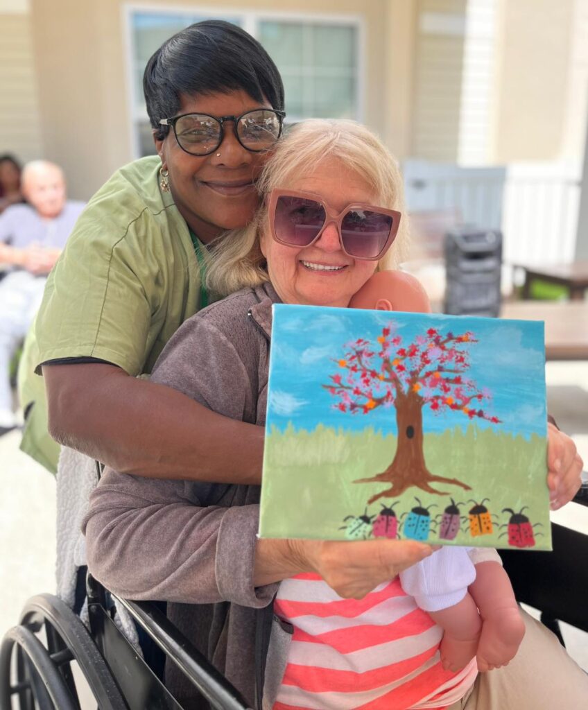 A resident with memory loss holding up a painting with a Village Green staff member embracing them