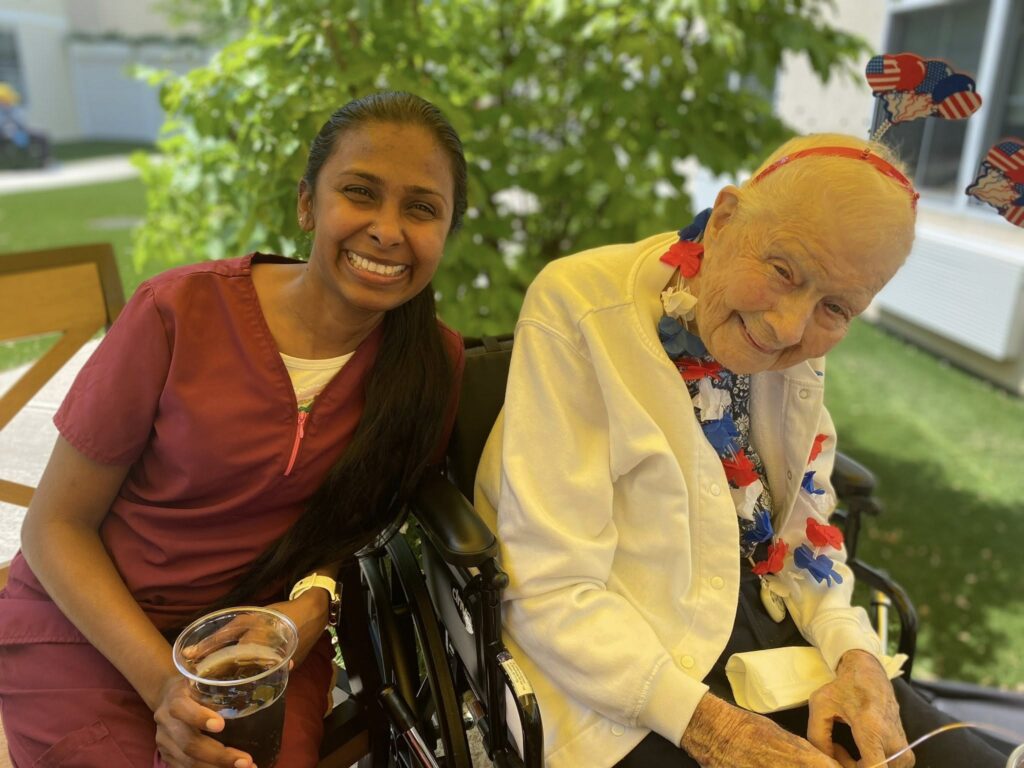 A senior citizen in a wheelchair smiling next to a registered nurse who is also smiling